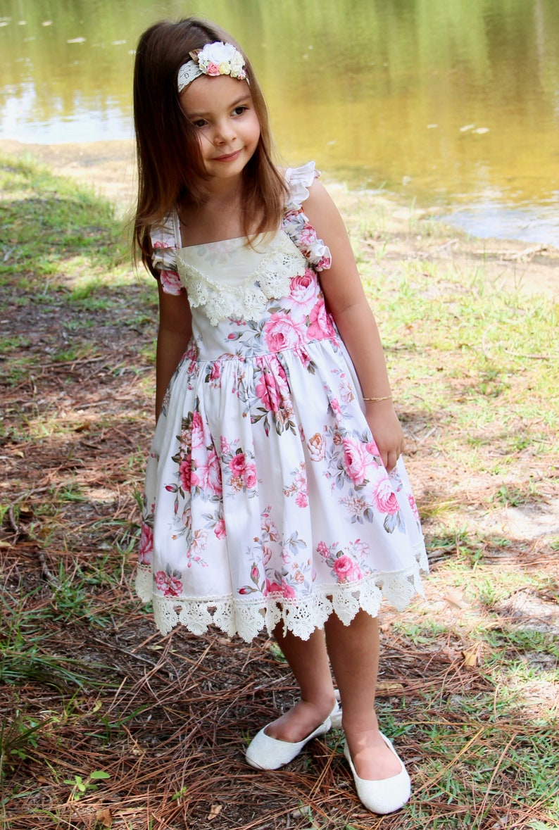 May include: A young girl wearing a white dress with pink and gray floral print and a white lace trim. She is wearing a white headband with a floral design and white shoes. The girl is standing in a grassy area with pine needles on the ground.