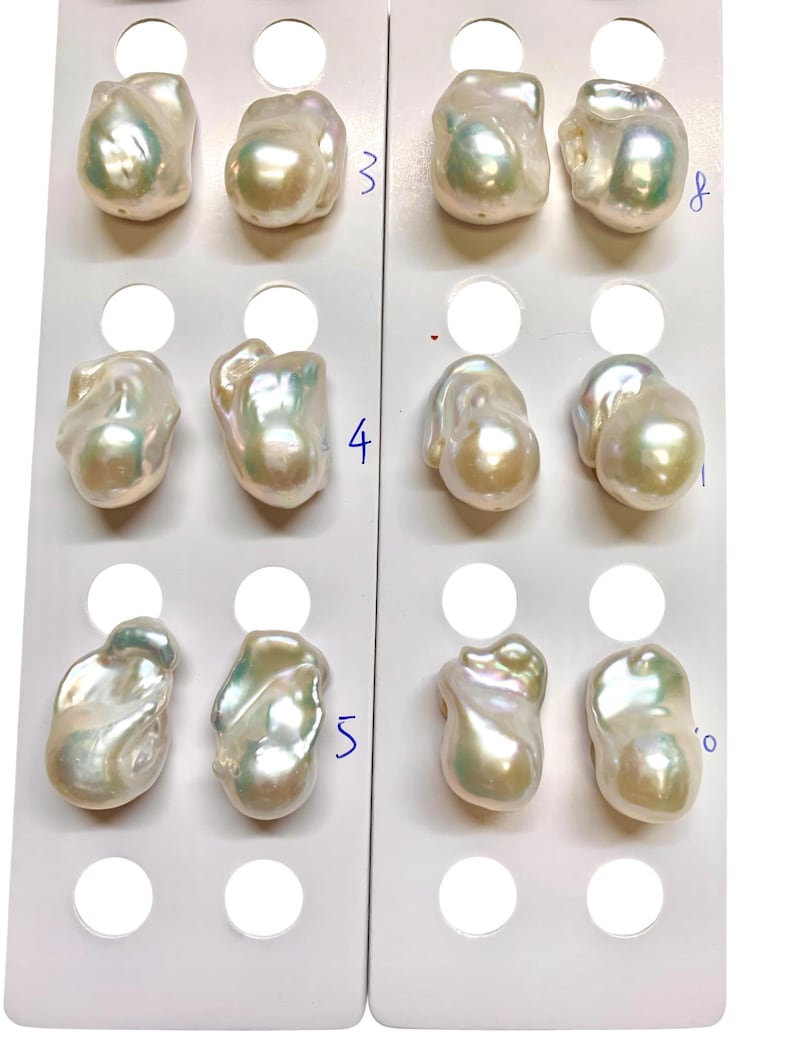 May include: A collection of lustrous, irregularly shaped pearls, each with a creamy white base and iridescent sheen. The pearls are arranged on white display cards, showcasing their unique forms and natural beauty. Numbers are visible on the cards.