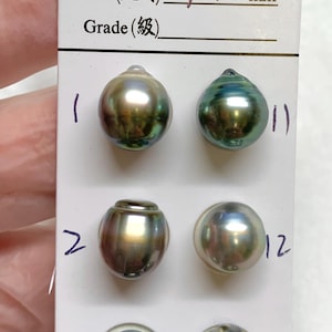 May include: A selection of Tahitian pearls in various shapes and colors, displayed on a white card. The card includes handwritten text indicating size as 9-10 mm. The pearls range from silver to dark green.