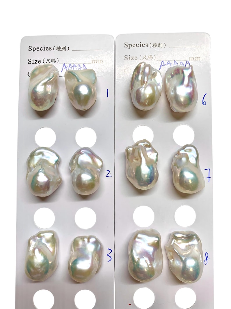 May include: Close-up of two white cards displaying irregularly shaped pearls. Each card has a series of pearls, numbered 1 through 8. The pearls have a lustrous, iridescent sheen, reflecting colors like pink, green, and gold.