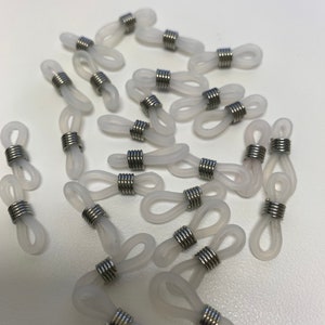 以下が含まれることがあります： 白いシリコン製の眼鏡コードホルダー25個と、銀色の金属リングがセットになっています。ホルダーは白い表面に散らばっています。