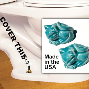 Puede incluir: Se muestran dos figuras de gato de cerámica turquesa, con un acabado brillante. Las figuras están etiquetadas como "Made in the USA". La imagen también muestra un inodoro blanco con el texto "COVER THIS".