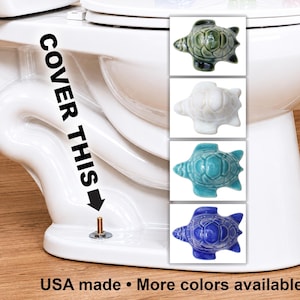 May include: White ceramic toilet with a selection of turtle-shaped toilet bolt covers in green, white, turquoise, and blue. The text "COVER THIS" is printed on the side of the toilet.