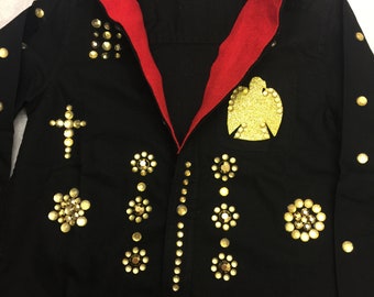 May include: A black denim shirt with a red lining and gold embellishments. The shirt has a gold cross on the left side and a gold eagle on the right side. The shirt is covered in gold studs.