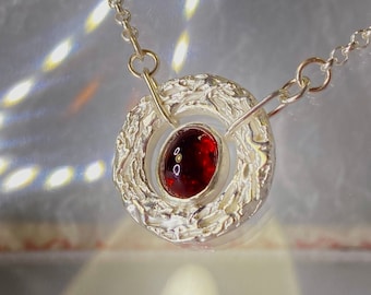 May include: A silver necklace with a pendant holding a red gemstone. The pendant has a circular, textured design around the gemstone. The chain is delicate silver. The background is a reflective surface, with light reflections.