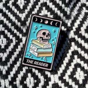 May include: A black enamel pin with a blue background and a white skull with a stack of books on top. The pin says "THE READER" in white letters.