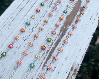 Puede incluir: Varios brazaletes de cadena de margaritas con acentos florales coloridos. Cada brazalete presenta una cadena plateada con pequeñas flores vibrantes en tonos rosa, amarillo, azul, verde y rojo. Se exhiben sobre una superficie de madera blanca envejecida.