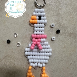 May include: A goose-shaped keychain crafted from white, orange, and pink plastic pony beads, featuring a silver metal ring at the top. The design includes a pink bead scarf and orange feet. A logo reads EVERLEE RAE OF Sunshine.