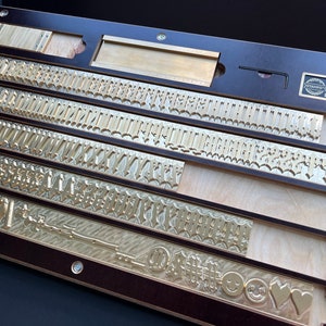 Könnte beinhalten: Ein Holztablett mit fünf Reihen von Messingbuchstaben. Die Stempel sind alphabetisch geordnet und umfassen Groß- und Kleinbuchstaben, Zahlen und Symbole. Das Tablett ist mit "Iron Horse Stamp Co. Sapporo, Japan." beschriftet.