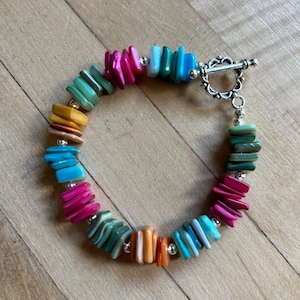 May include: A bracelet featuring multi-colored, rectangular shell chip beads in shades of pink, turquoise, yellow, orange, and blue, interspersed with small silver-toned metal beads and secured with a silver-toned metal toggle clasp closure.