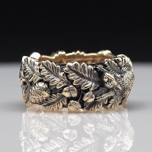 May include: A gold and silver ring with a detailed design of oak leaves, acorns, and a bird. The ring is wide and has a textured surface.
