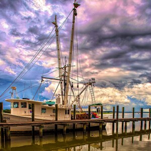 May include: A white fishing boat named "Miss Joyce" is docked at a wooden pier. The boat has a tall mast and a large net hanging from it. The sky is cloudy and dramatic, with shades of purple, pink, and gray.