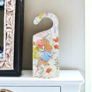May include: A wooden doorstop shaped like a hanger with a blue and white floral pattern. The doorstop features a Peter Rabbit illustration with a red ladybug.
