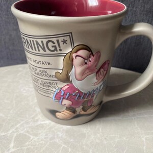 Puede incluir: Una taza de cerámica beige con interior rojo intenso que presenta un gráfico del personaje Gruñón. El texto dice WARNING! DO NOT AGITATE. DO NOT ASK QUESTIONS. DO NOT EXPECT A HAPPY RESPONSE. La palabra Grumpy está impresa en azul.