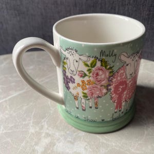 Peut inclure: Une tasse en céramique avec un fond vert clair à pois blancs, illustrée de deux moutons. L'un est orné de fleurs roses et violettes, l'autre porte un ruban rose avec l'inscription 3rd. Les textes Molly et best in show sont imprimés sur la tasse.