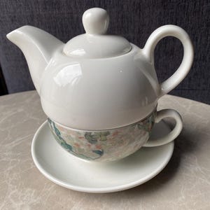 May include: A white ceramic tea set comprising a teapot with a lid, a teacup, and a saucer. The teacup has a floral pattern in green, blue, and beige. The set is designed for a single serving of tea.