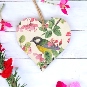 Może przedstawiać: Drewniana ozdoba w kształcie serca z kwiatowym wzorem i ptakiem. Serce jest pomalowane na biało i przedstawia różowe kwiaty, zielone liście i żółto-niebieskiego ptaka.