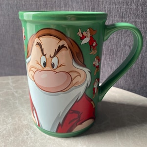 May include: A green ceramic mug featuring a large, detailed illustration of the character Grumpy from Snow White and the Seven Dwarfs. The mug has a glossy finish, a curved handle, and smaller repeated images of the same character in various poses.
