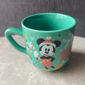 Może przedstawiać: Turkusowy ceramiczny kubek z grafiką przedstawiającą Myszkę Miki w czerwono-białej czapce Mikołaja, trzymającą czerwono-biały cukierek w kształcie laski. Wzór zawiera świąteczne elementy, takie jak zielona choinka oraz białe gwiazdki.