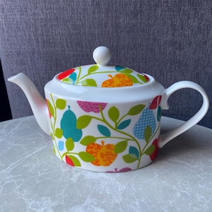 Op de afbeelding: Witte keramische theepot met deksel, tuit en handvat. De theepot is versierd met een kleurrijk patroon van fruit en bladeren in tinten rood, oranje, blauw en groen. Het deksel heeft een kleine witte knop.