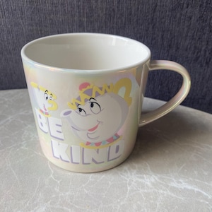 May include: Iridescent white ceramic mug featuring a cartoon teapot and teacup design. The mug has the words "BE KIND" printed on the front. The handle and the rim of the mug have a pearlescent finish.