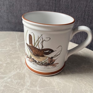 Może przedstawiać: Biały ceramiczny kubek ze szczegółową ilustracją brązowego strzyżyka siedzącego wśród zielonych traw i opadłych liści. Kubek posiada gładkie ucho oraz charakterystyczną czerwonobrązową obwódkę wokół górnej krawędzi i podstawy naczynia.
