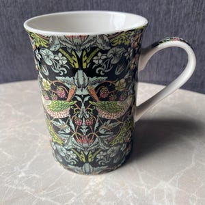 Puede incluir: Una taza de cerámica con interior y asa blancos. El exterior presenta un detallado estampado floral y de pájaros en tonos verdes, rosas y crema sobre un fondo negro. La taza está diseñada para bebidas calientes.