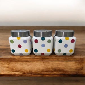 May include: Three white ceramic jars with stainless steel lids. Each jar is decorated with colourful polka dots in red, green, yellow, blue, and purple. The jars are square-shaped with rounded corners and sit on a wooden surface.