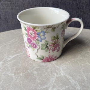 Può includere: Una tazza in ceramica bianca con un motivo floreale con fiori rosa, blu e verdi. La tazza ha un manico curvo e un bordo leggermente svasato. Il motivo floreale è su uno sfondo color crema.