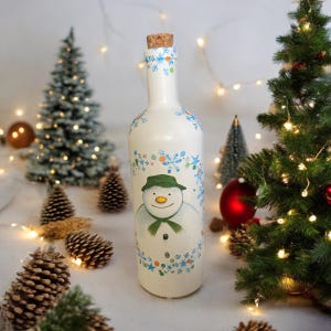 Puede incluir: Una botella de vidrio blanco con un tapón de corcho está decorada con un diseño de muñeco de nieve. El muñeco de nieve lleva un sombrero verde y una bufanda verde. La botella está rodeada de piñas y un pequeño árbol de Navidad.