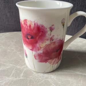Könnte beinhalten: Ein weißer Keramikbecher mit einem floralen Aquarell-Design, das leuchtend rote und rosafarbene Mohnblumen, grüne Stängel und kleine Knospen zeigt. Der Becher hat einen glatten, zylindrischen Körper mit einem geschwungenen Henkel.