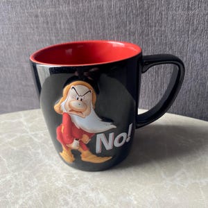 May include: A black ceramic mug with a bright red interior and a black handle. The exterior features a raised, three-dimensional graphic of the character Grumpy from Snow White, wearing a red tunic and yellow shoes, with the word No! printed in white.