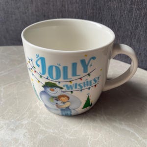Może przedstawiać: Biały ceramiczny kubek z niebieskim napisem JOLLY WISHES! nad ilustracją bałwana w zielonym kapeluszu i szaliku, trzymającego małe dziecko. Wzór zawiera kolorowe lampki choinkowe oraz małe zielone choinki świąteczne.