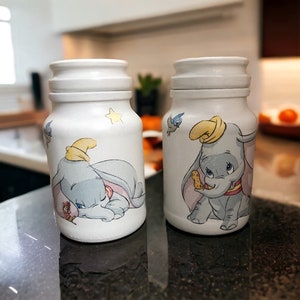 May include: Two white ceramic jars decorated with a cartoon image of Dumbo the elephant. One jar features Dumbo sleeping with a star above him, while the other jar shows Dumbo sitting up with a peanut in his trunk.