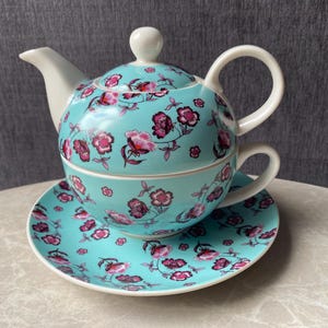Könnte beinhalten: Türkisfarbenes Keramik-Teeservice mit floralem Muster. Das Set beinhaltet eine Teekanne, eine Tasse und eine Untertasse. Das florale Design zeigt rosa und lila Blumen mit grünen Stielen. Die Teekanne hat einen weißen Griff und Ausguss.