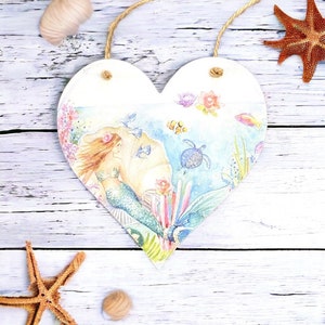 Puede incluir: Un letrero de madera en forma de corazón con una imagen pintada de una sirena nadando en un arrecife de coral. La sirena tiene el pelo largo y rojo que fluye y una cola verde. El arrecife está lleno de peces coloridos y una tortuga marina.
