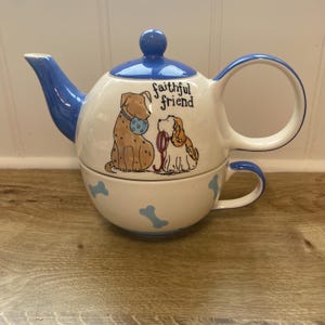 May include: A white ceramic teapot and teacup set with blue accents. The teapot features a blue lid and spout, and a large handle. The teacup has a blue handle and bone-shaped designs. The set is decorated with illustrations of two dogs and the text "faithful friend."
