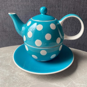 Może przedstawiać: Turkusowy ceramiczny zestaw do herbaty składający się z czajniczka z pokrywką i pasującej filiżanki, oba ozdobione wzorem w białe kropki. Czajniczek ma zakrzywiony uchwyt i dzióbek, a filiżanka stoi na pasującym turkusowym spodku.