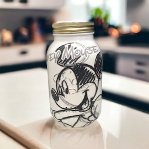 Op de afbeelding: Een witte glazen pot met een gouden deksel waarop een zwart-witte schets van Mickey Mouse te zien is. De pot heeft de tekst "Mickey Mouse" erop.