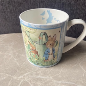 May include: White ceramic mug featuring a Peter Rabbit design. The mug has a light blue border with illustrations of rabbits and butterflies. The main image depicts Peter Rabbit in a blue jacket near a watering can.