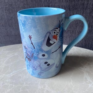 May include: A light blue ceramic mug featuring a cartoon image of Olaf from the movie Frozen. The mug has a light blue interior and handle. The exterior has a wintery design with snowflakes and ice.