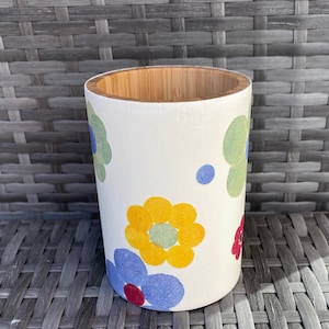 May include: A white bamboo pencil holder with a floral design. The design features blue, yellow, green, and red flowers.