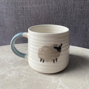 May include: A cream-coloured ceramic mug with a ribbed texture and a light blue handle. The front of the mug features a printed illustration of a fluffy sheep with a black face and legs, rendered in a simple, clean style on the rounded body.