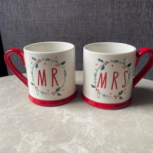 May include: Two cream-coloured ceramic mugs with red handles and red bases. Each mug features a green holly leaf and red berry wreath design. One mug has the red text MR in the centre, and the other has the red text MRS in the centre.