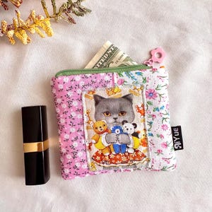 May include: A small, square coin purse with a floral pattern and a cat illustration holding teddy bears. The purse has a green zipper and a pink flower-shaped zipper pull. A black lipstick tube is next to the purse.