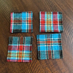 May include: Four square fabric coasters with a plaid pattern. The coasters feature a mix of colors, including blue, red, yellow, and black. The coasters are arranged on a wooden surface.