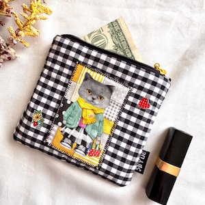 May include: A square black and white gingham patterned coin purse with a zipper closure. The purse features a patch with a cartoon cat wearing a scarf and coat. A lipstick and a dollar bill are next to the purse.