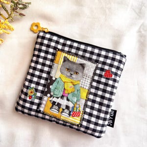 May include: A square black and white gingham patterned zippered pouch. The pouch features a patch with a cartoon cat wearing a green jacket and yellow scarf. A small red heart and a flower are on the pouch.
