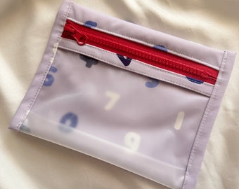 May include: A small, clear, lavender zippered pouch with a red zipper. The pouch has a white background with a repeating pattern of blue and white numbers.