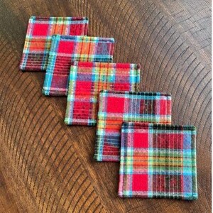 May include: Five square coasters with a plaid pattern in red, orange, blue, and green. The coasters are arranged diagonally on a wooden surface, showcasing their colorful design and texture. These coasters are designed to protect surfaces from drink rings.